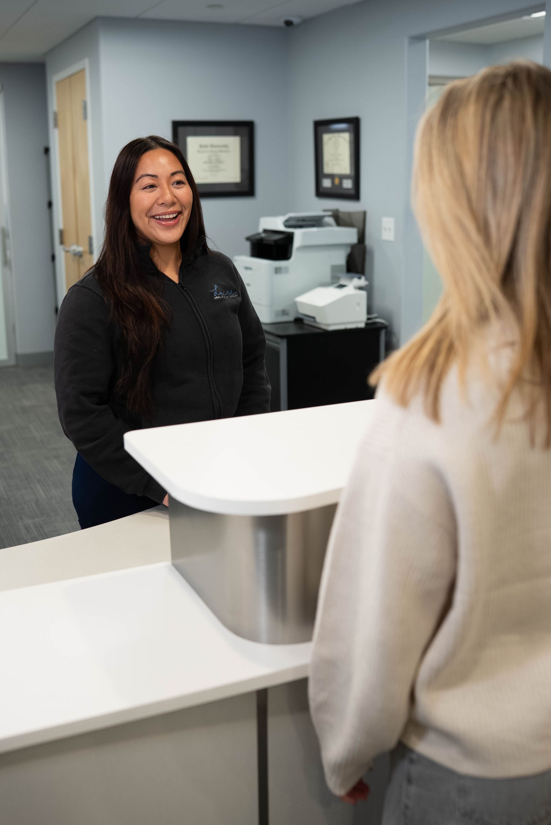 A person at a reception desk hands paperwork to a smiling staff member in a gray zip-up jacket in an office setting.