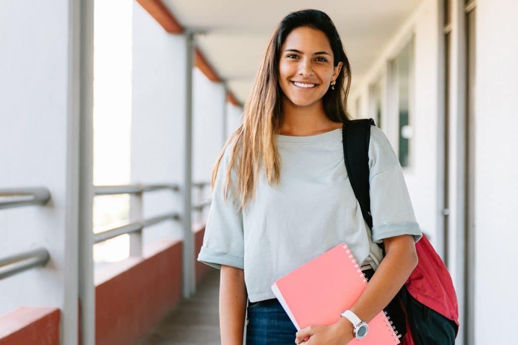 A smiling student holding a pink notebook, wearing a backpack, standing in a brightly lit school corridor.