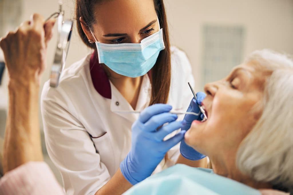 A dentist in a mask and blue gloves examines a patient's teeth while the patient holds a mirror.