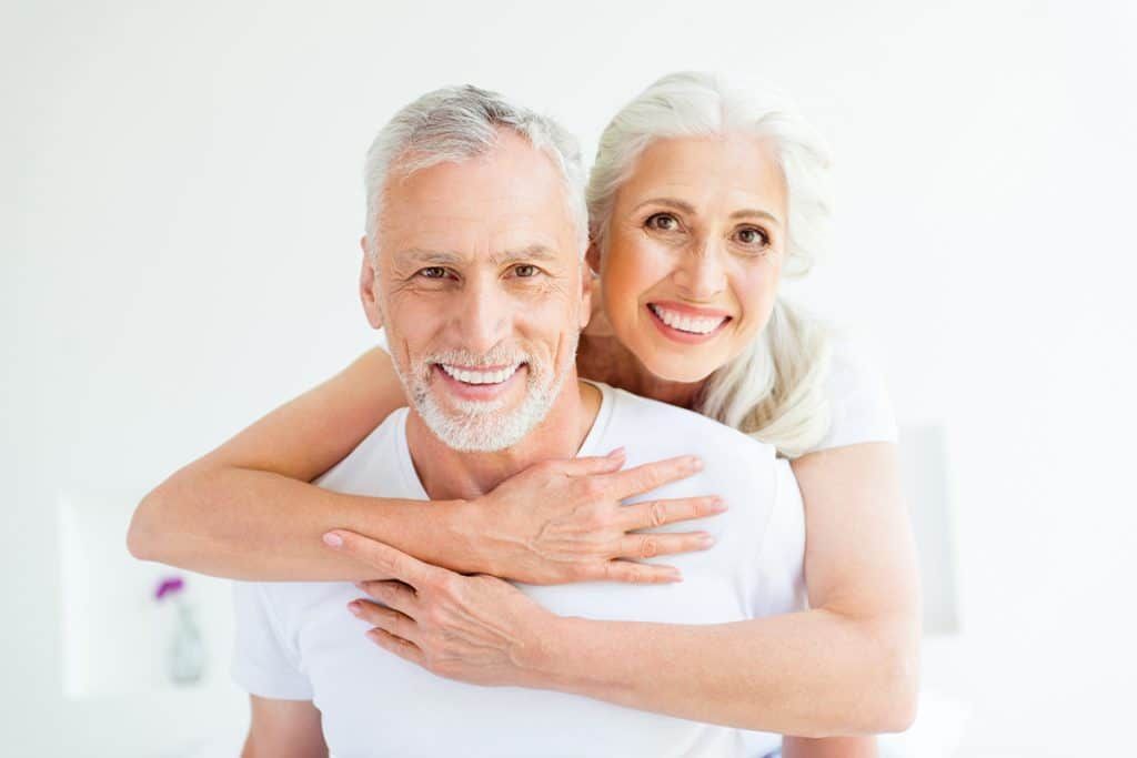 A smiling person with gray hair hugs another person from behind against a plain white background.