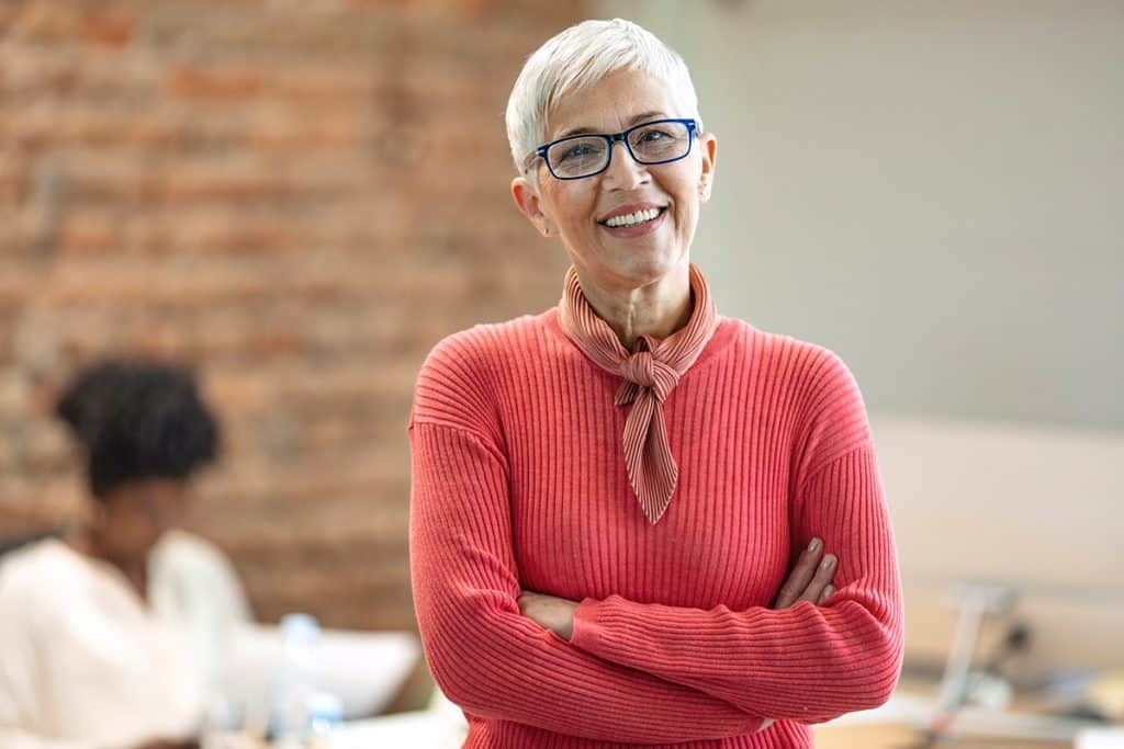 A smiling person wearing a coral-colored textured sweater and glasses stands with arms crossed in an office.