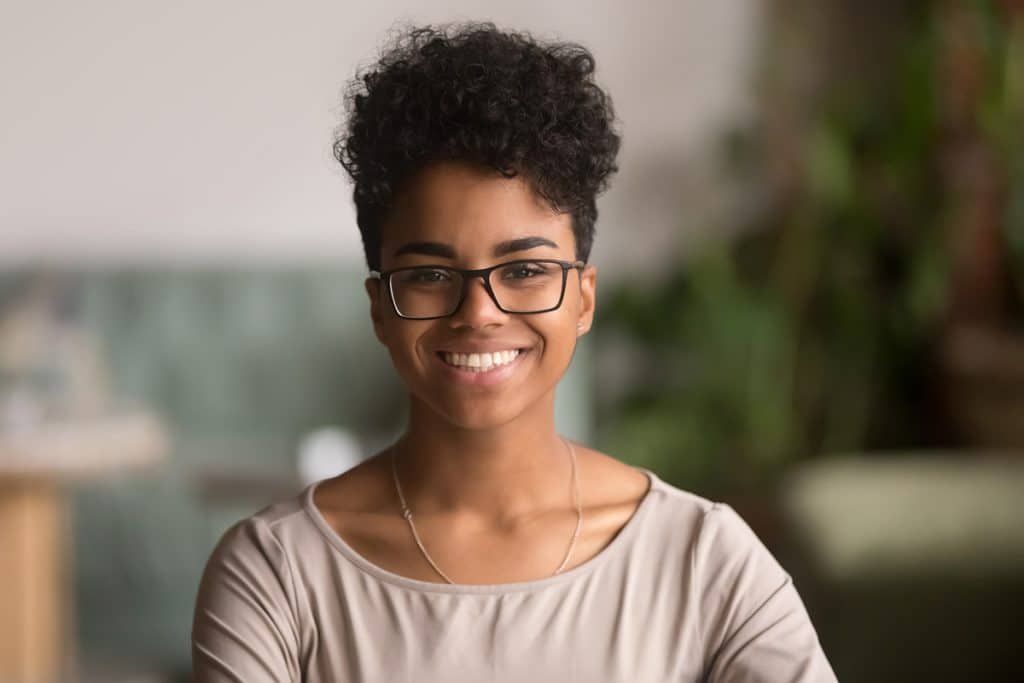 A person with short, curly hair and glasses smiles while wearing a light-colored top in a softly lit, blurred interior.
