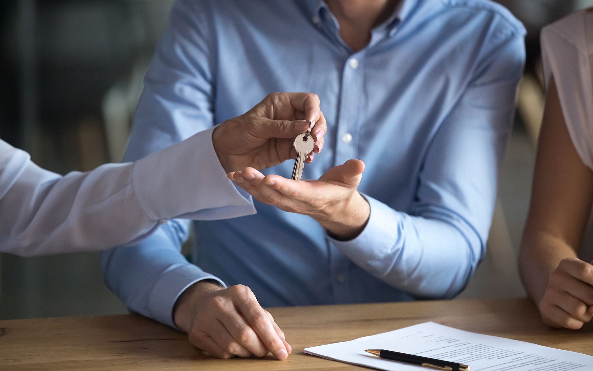 Person handing keys to another person over a document on a wooden table.
