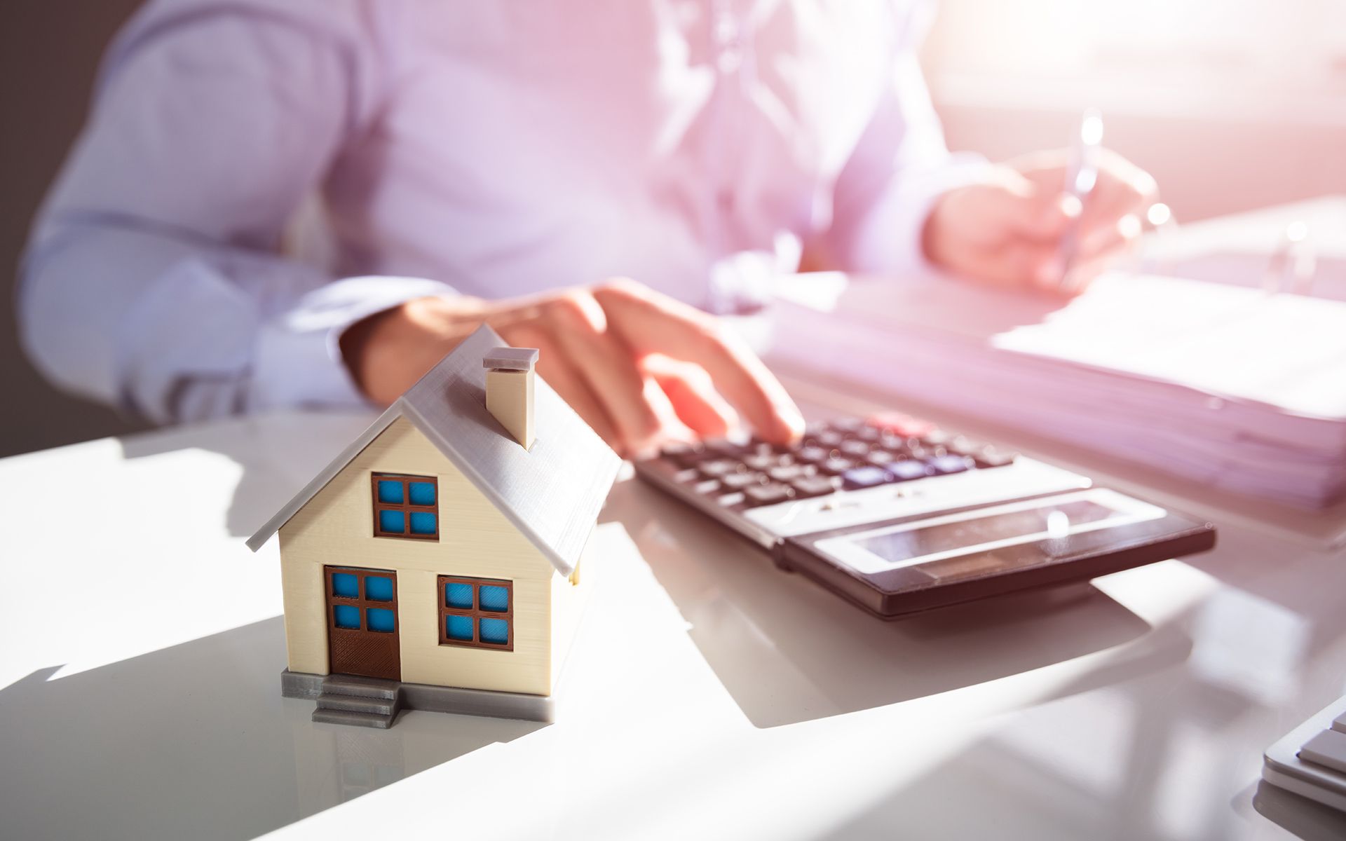 A person calculates with a calculator next to a toy house on a desk with paperwork.
