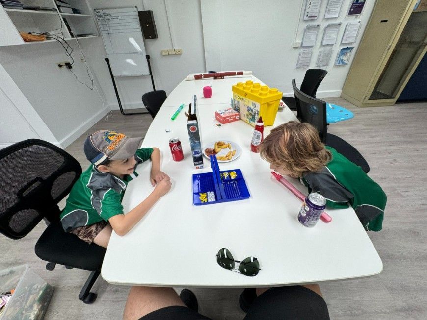 Two people sit across from each other at a table in a room, focusing on a blue board game, with drinks and snacks nearby — Specialist Support Solutions in Rockhampton City, QLD