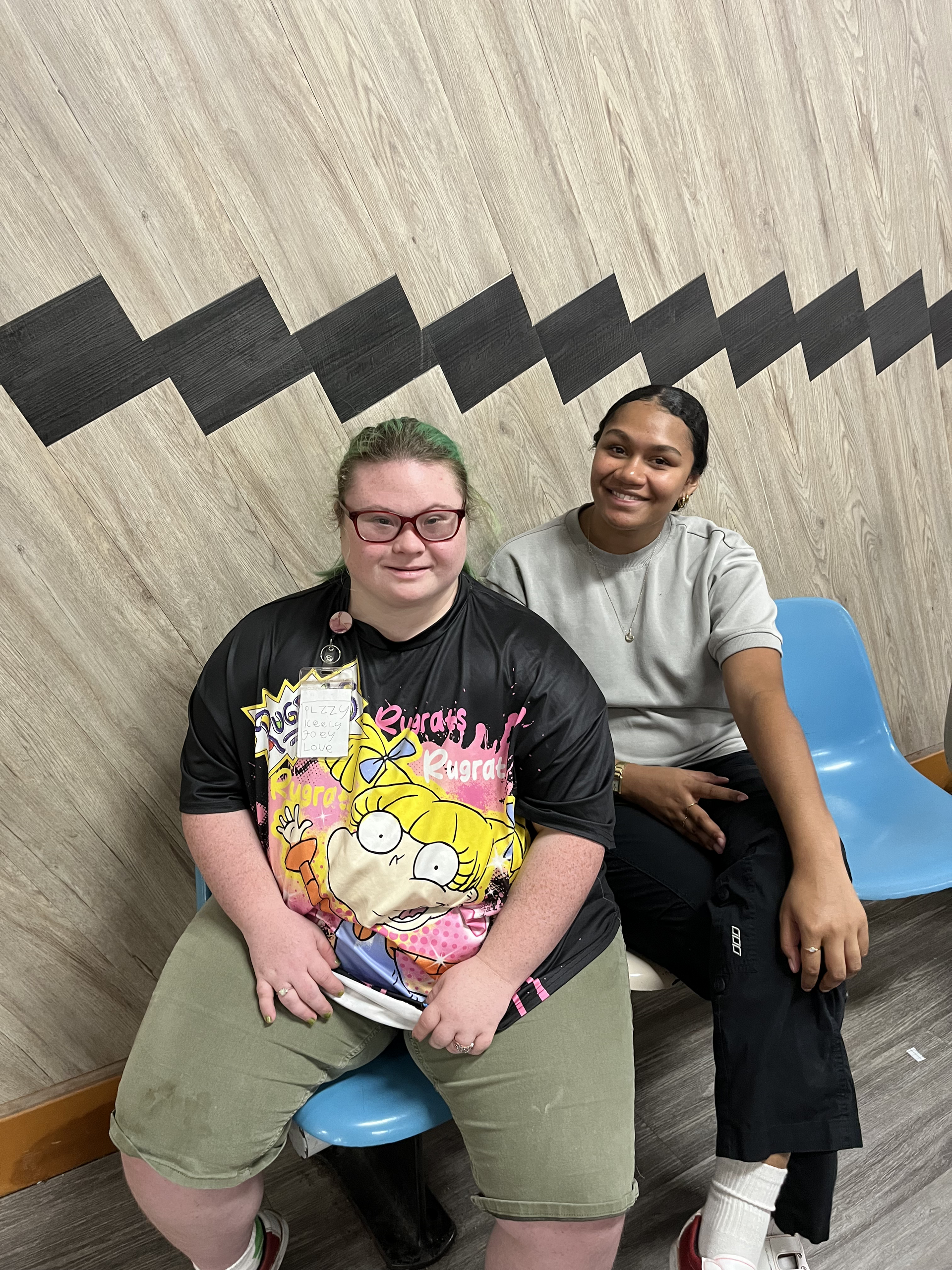 Two people sit side-by-side on blue chairs against a wall with a black geometric pattern, smiling at the camera — Specialist Support Solutions in Rockhampton City, QLD
