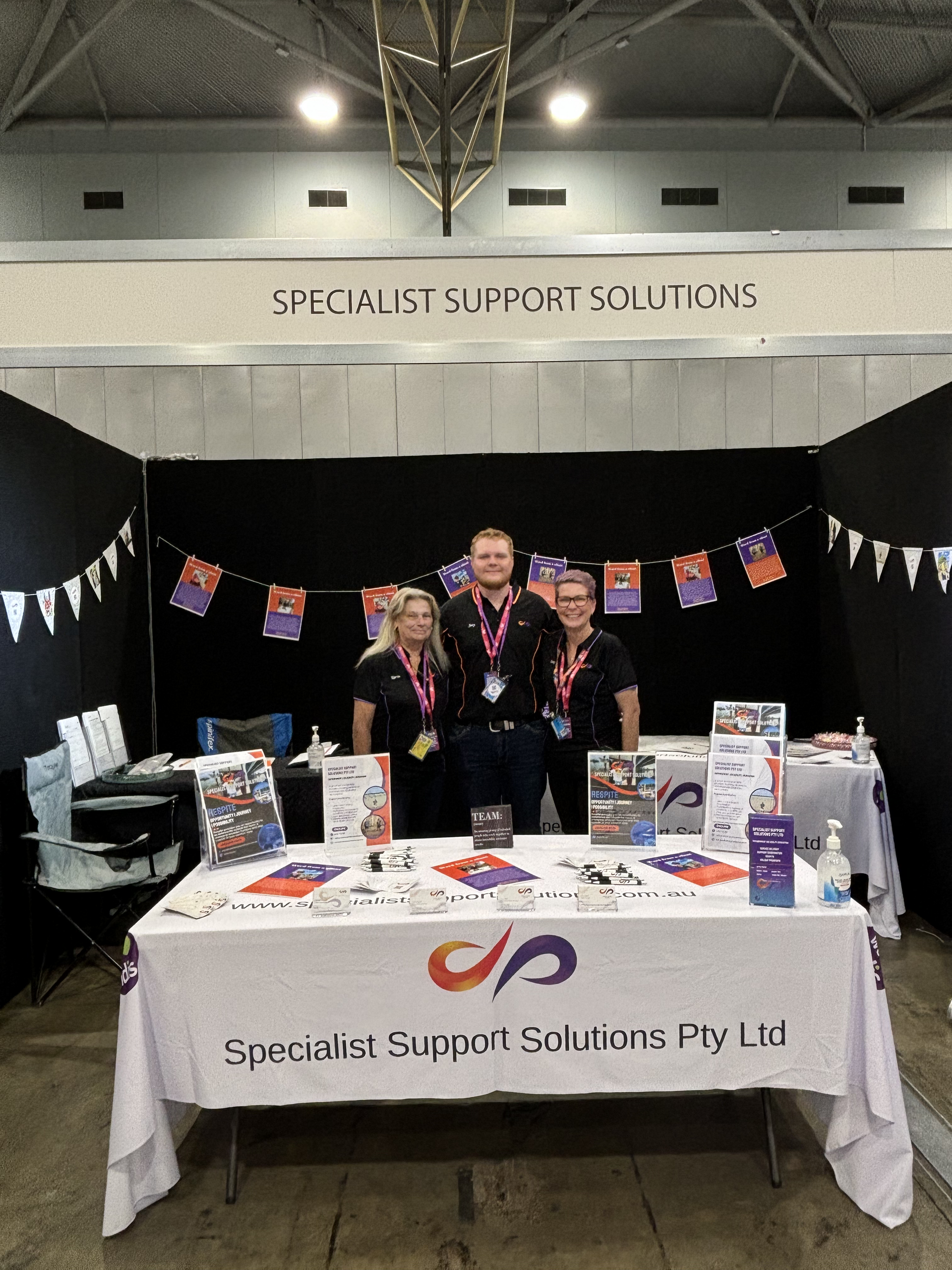 Three people stand behind a branded Specialist Support Solutions booth at an event, with brochures on a white table -— Specialist Support Solutions in Rockhampton City, QLD
