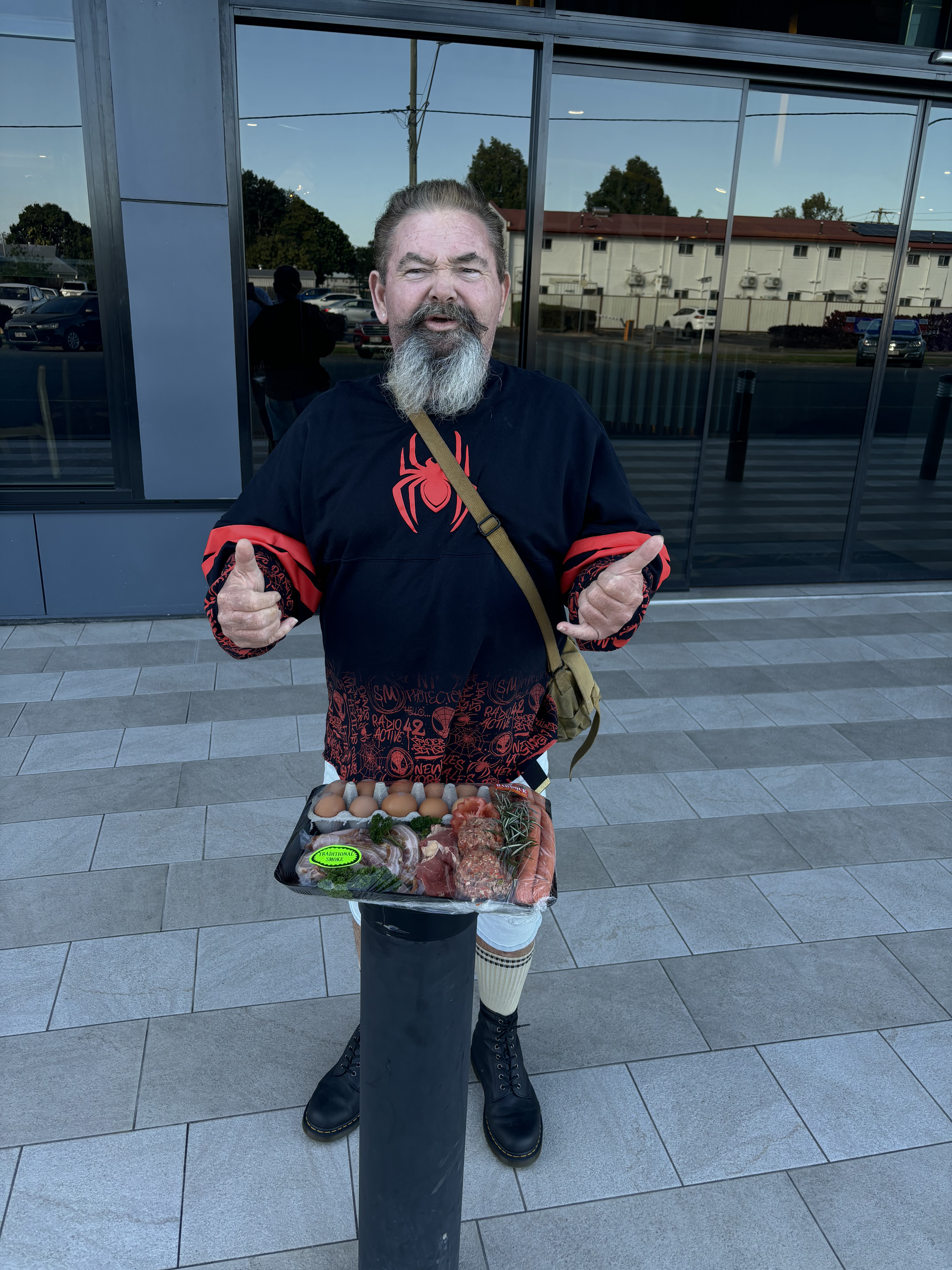 A person with a beard and a spider-themed sweater gives a thumbs-up while holding a tray of food outdoors — Specialist Support Solutions in Rockhampton City, QLD