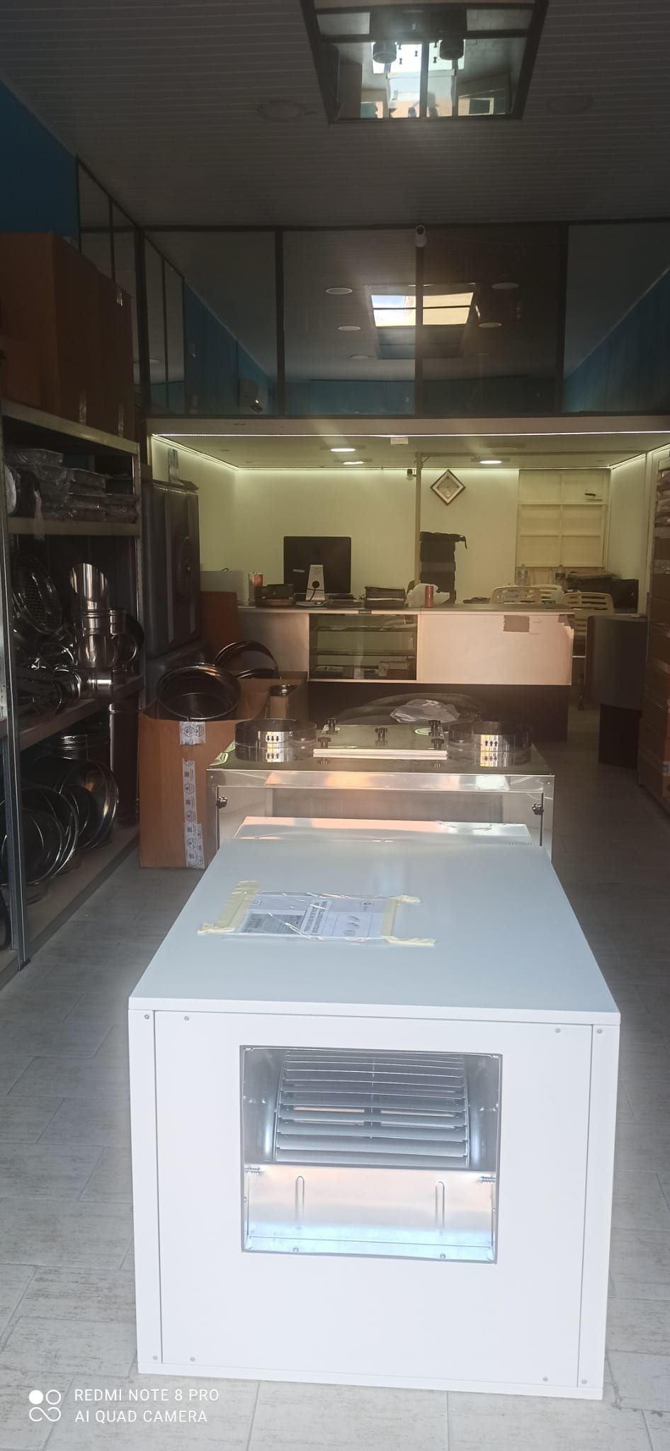 Vista frontale di un elettrodomestico bianco in una hall d'ufficio illuminata, con porte a vetri e scrivanie sullo sfondo.