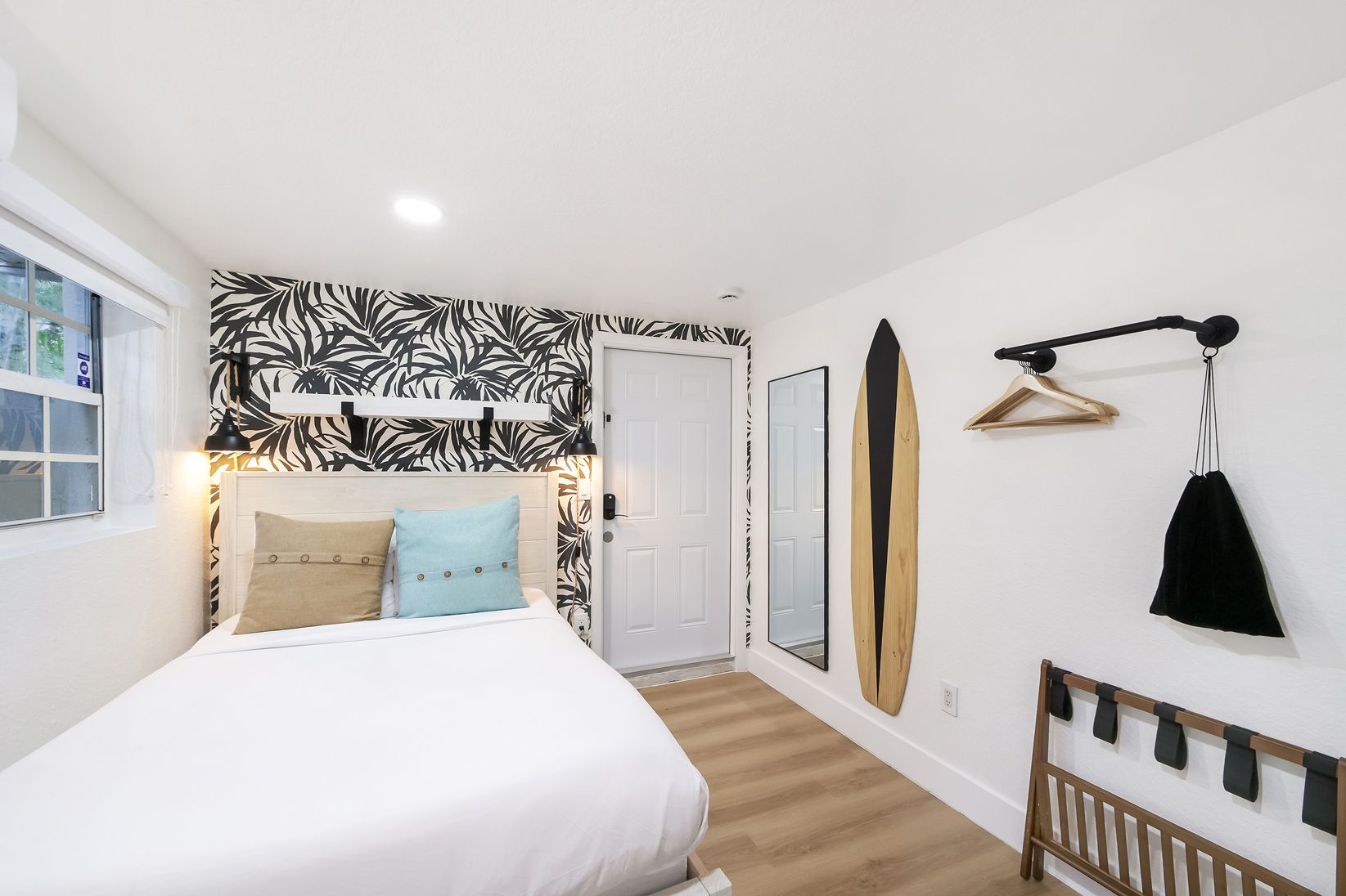 A bedroom with a queen bed, patterned wallpaper, a surfboard mounted on the wall, and a minimalist coat rack.
