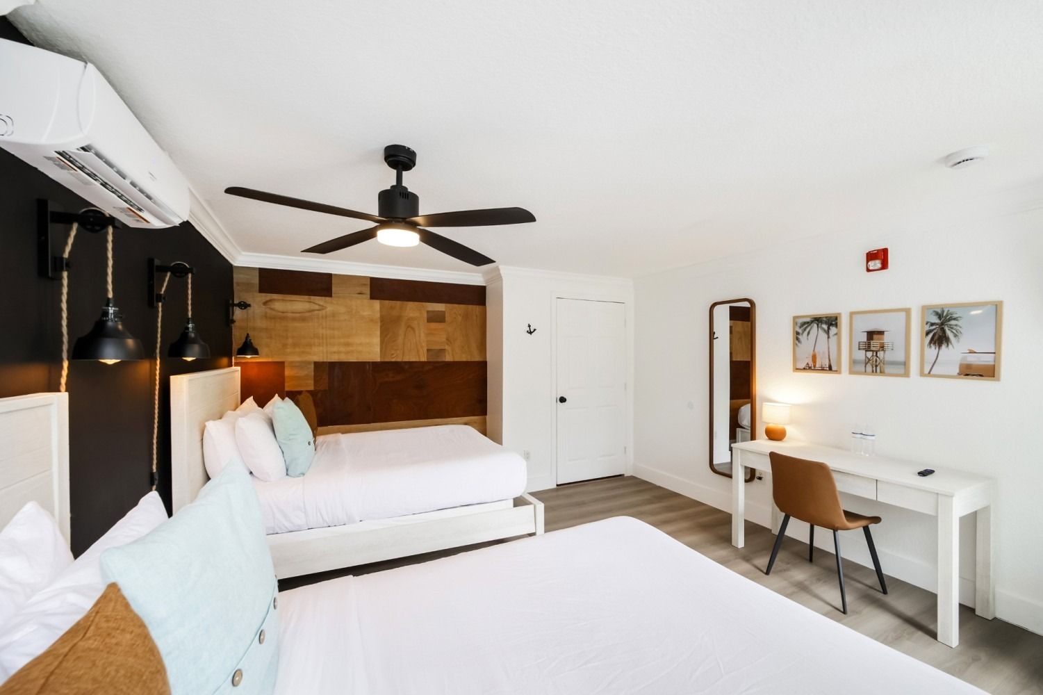 A hotel room with two white beds, a dark accent wall, a ceiling fan, a desk with a chair, and framed coastal wall art.