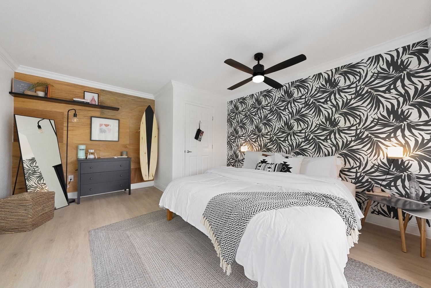 A modern bedroom with a black-and-white leaf wallpaper accent wall, wood-paneled corner, surfboard, and neutral bedding.
