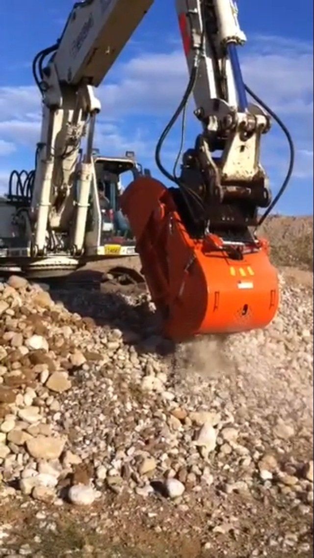 Un escavatore arancione frantuma rocce in un cantiere edile sotto un cielo azzurro.