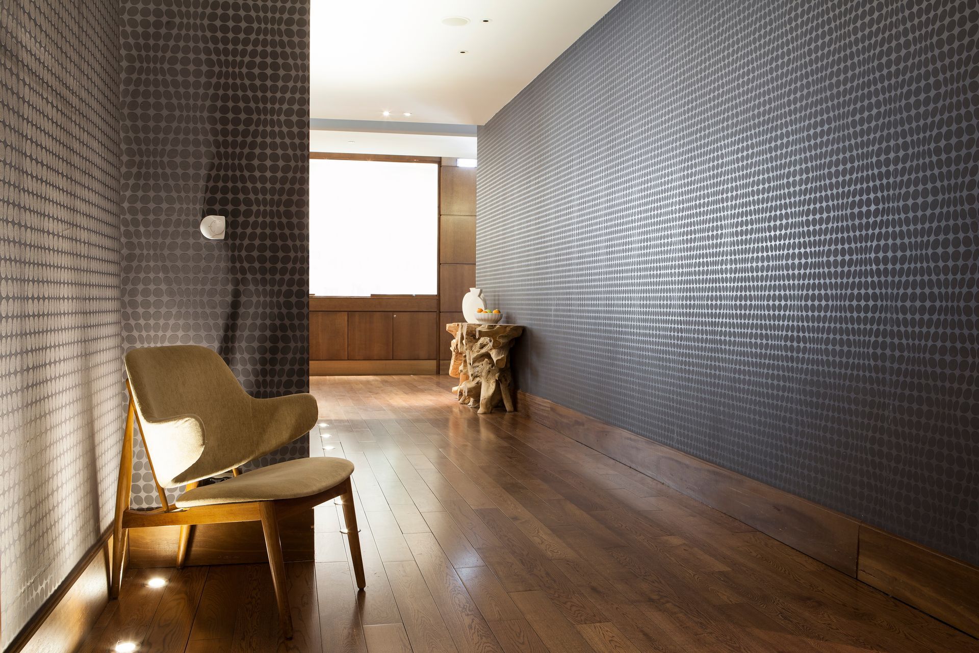 Long hallway with dark textured walls, wooden floor, and a chair by a bright window at the end.