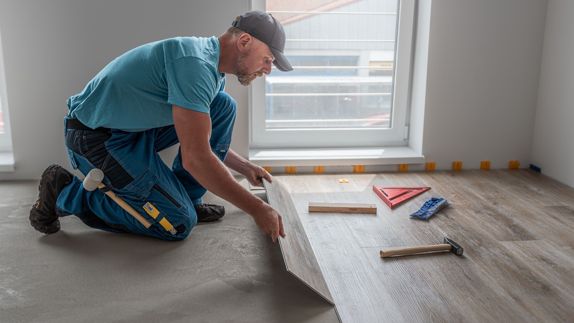 Worker kneeling on floor, installing gray carpet tiles beside tools and a window. Worker kneeling on floor, installing gray carpet tiles beside tools and a window.