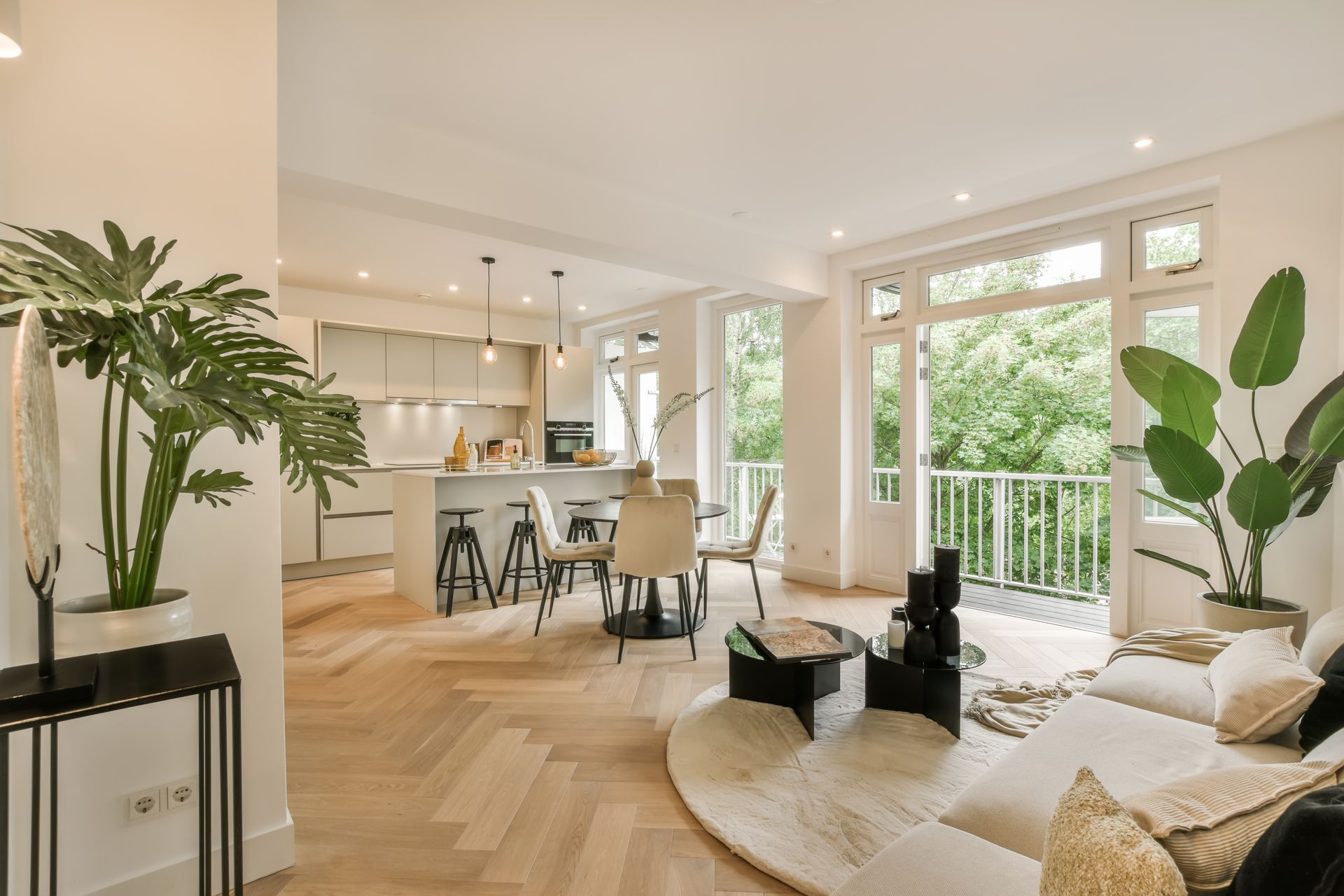 Bright open-plan living room with kitchen, herringbone floor, white sofa, plants, and balcony view Bright open-plan living room with kitchen, herringbone floor, white sofa, plants, and balcony view