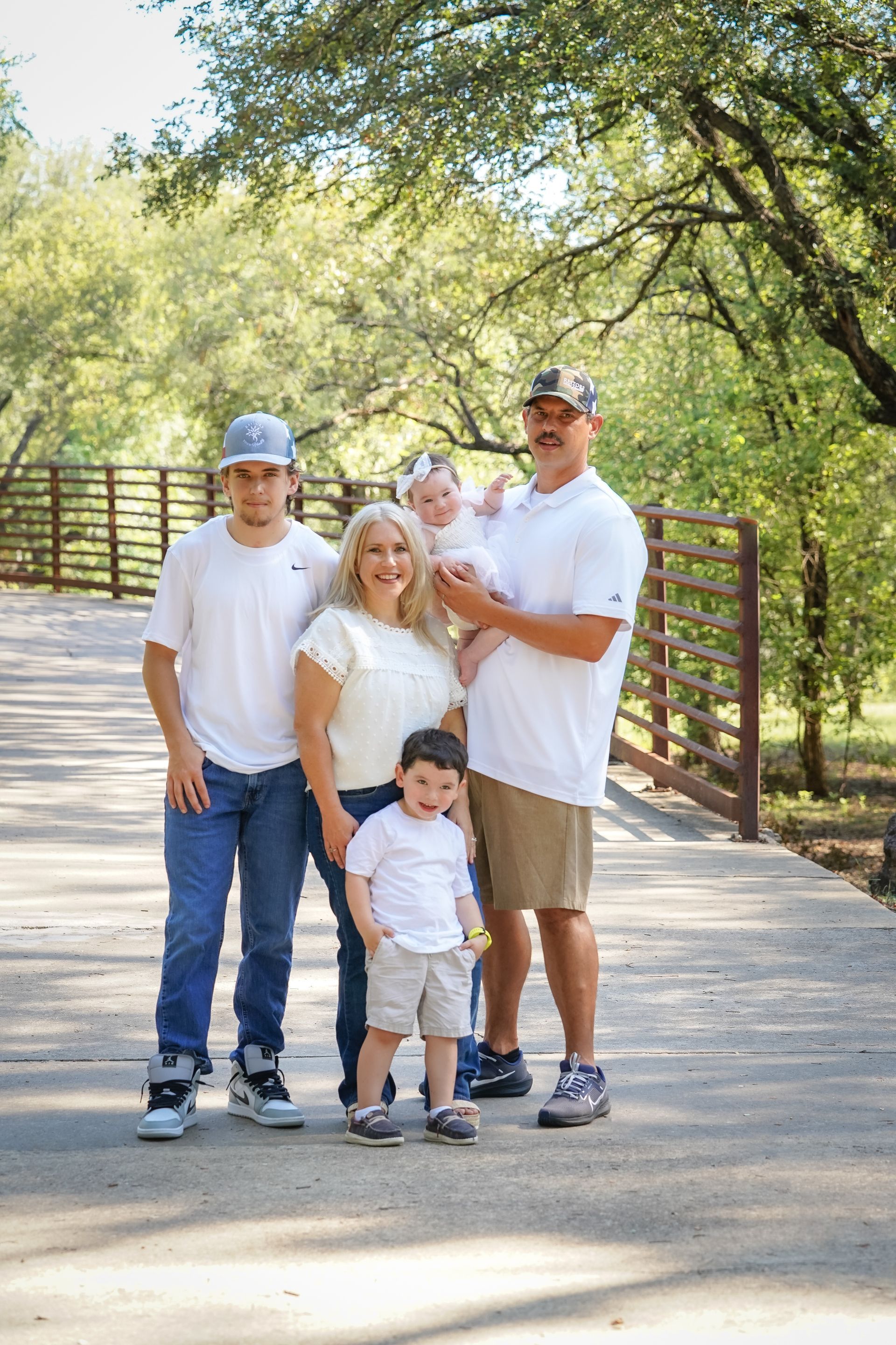 Family of five, posing on a bridge, under a tree. They wear white shirts and jeans/shorts.