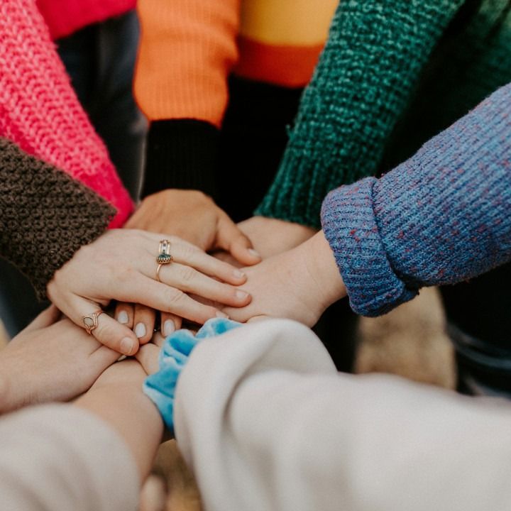 Hands stacked together in a circle, representing teamwork. Various colorful sweaters visible, outdoors.