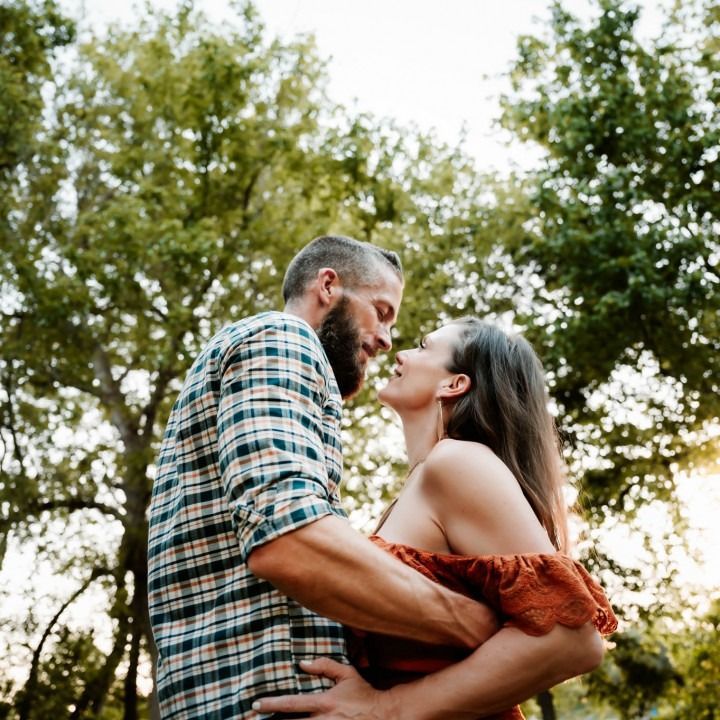 Couple embraces outdoors, faces close, woman in orange top, man in plaid shirt, green trees in background.