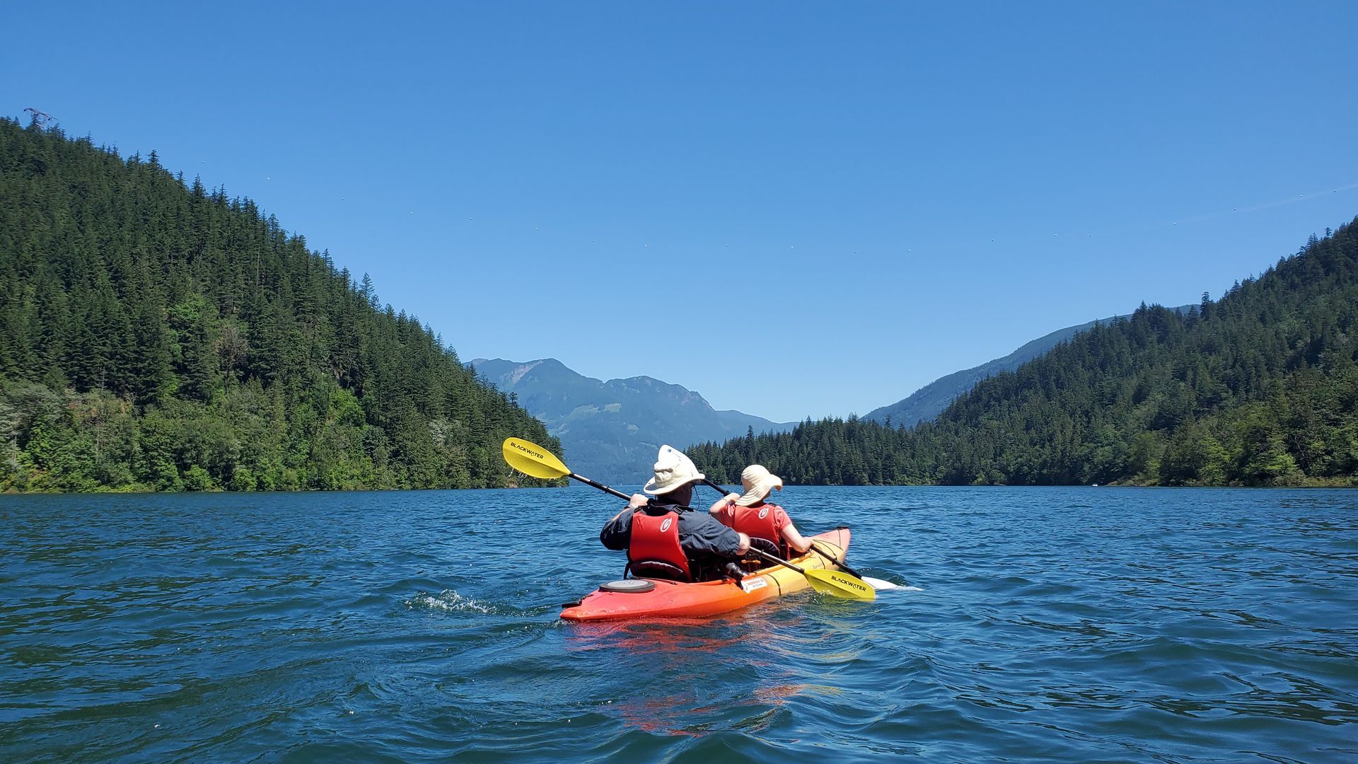 Two kayakers paddling on a blue lake between forested mountains under a clear sky
