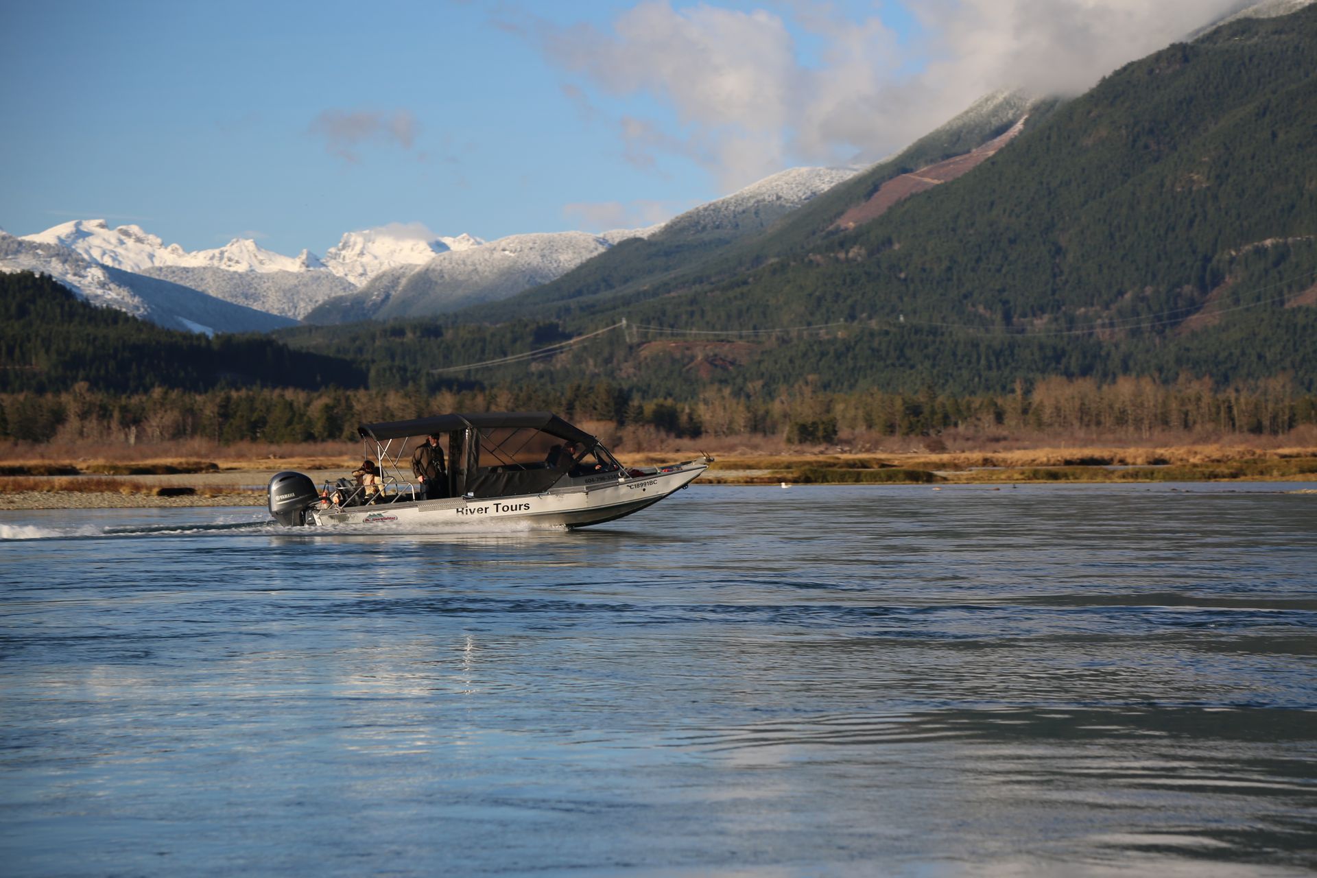Small motorboat on a river with snowy mountains and forested shoreline in the background