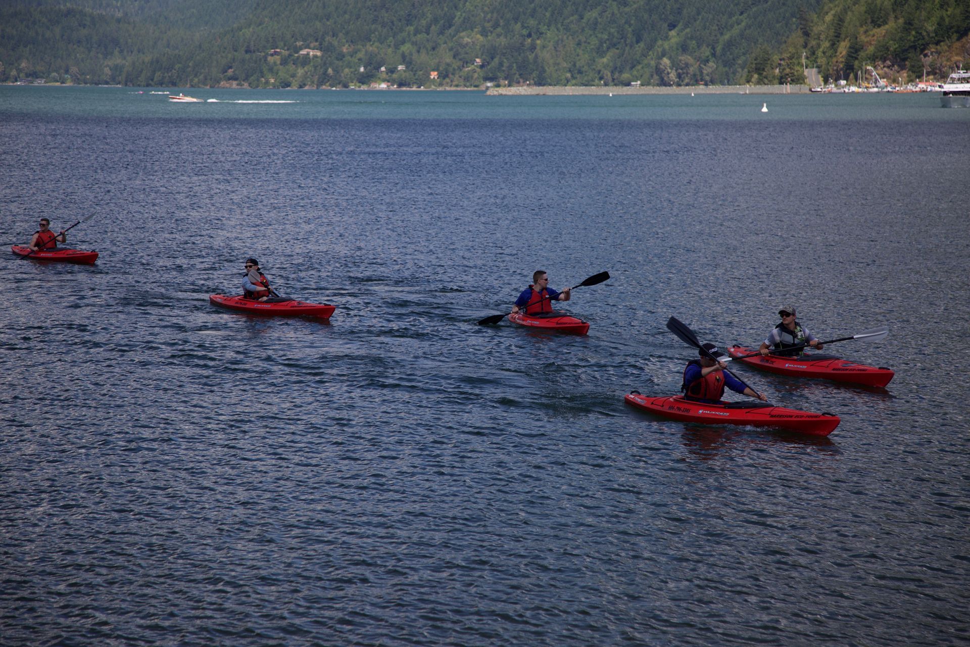 Kayakers in red boats paddling across a lake with forested hills in the background