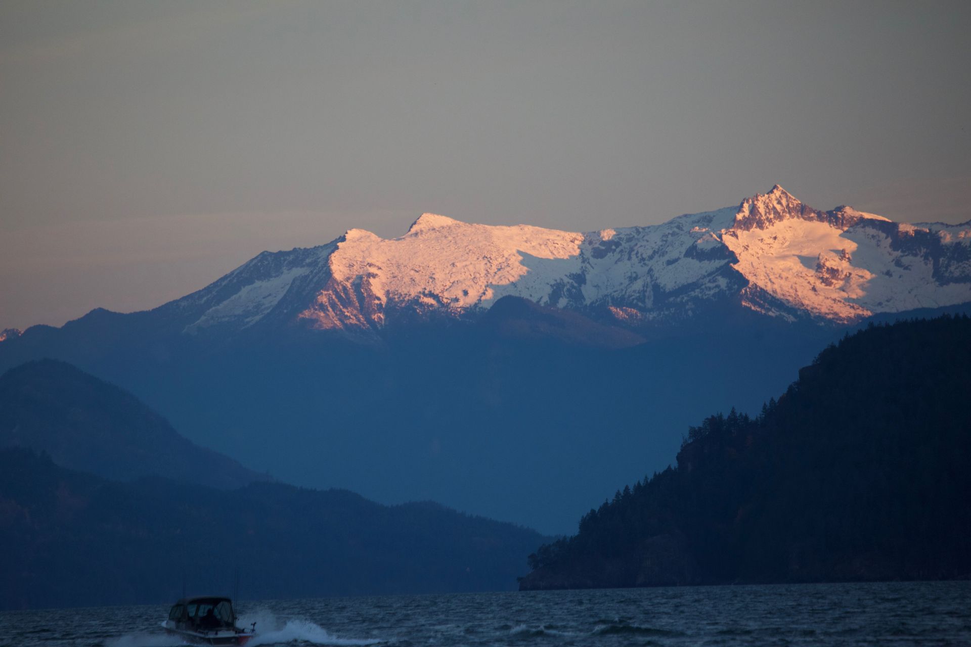 Snow-capped mountains at dawn, with pink-lit peaks above dark forested hills and a calm lake.