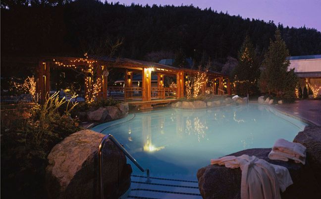 Illuminated outdoor hot spring pool at dusk beside a lit wooden lodge and snowy rocks