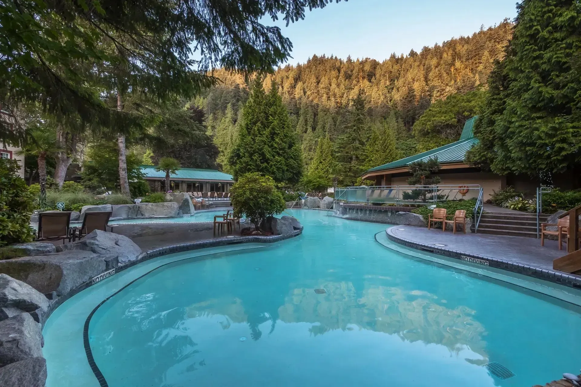 Outdoor hot spring pool with turquoise water, stone edges, and forested mountains in the background