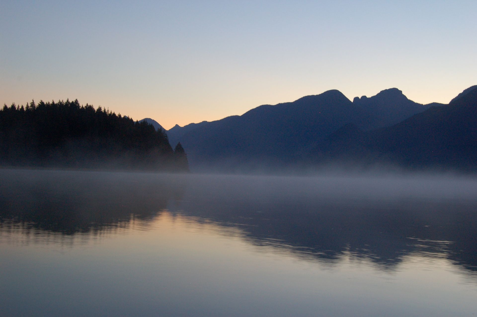 Calm lake at dawn with mountain silhouettes, mist, and reflections on the water