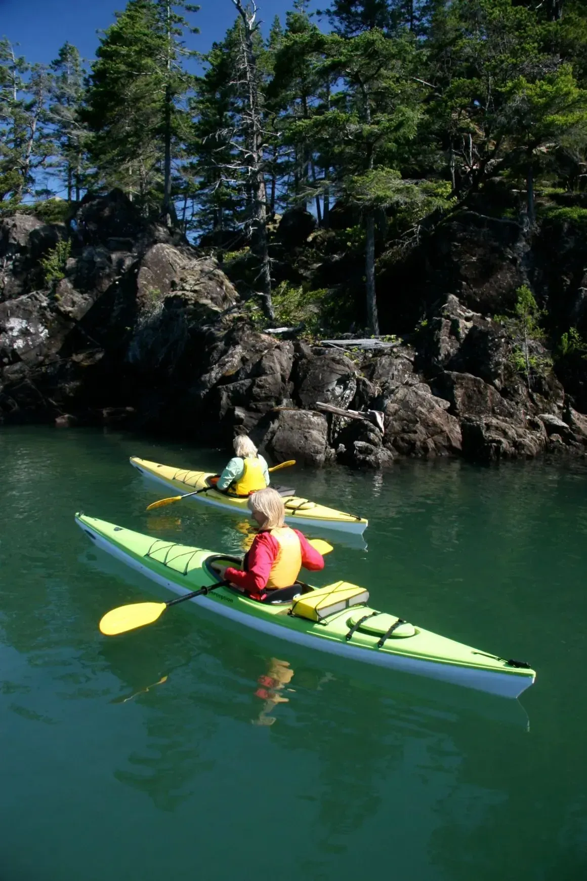 Two kayakers in yellow kayaks on green water near rocky, tree-lined shore