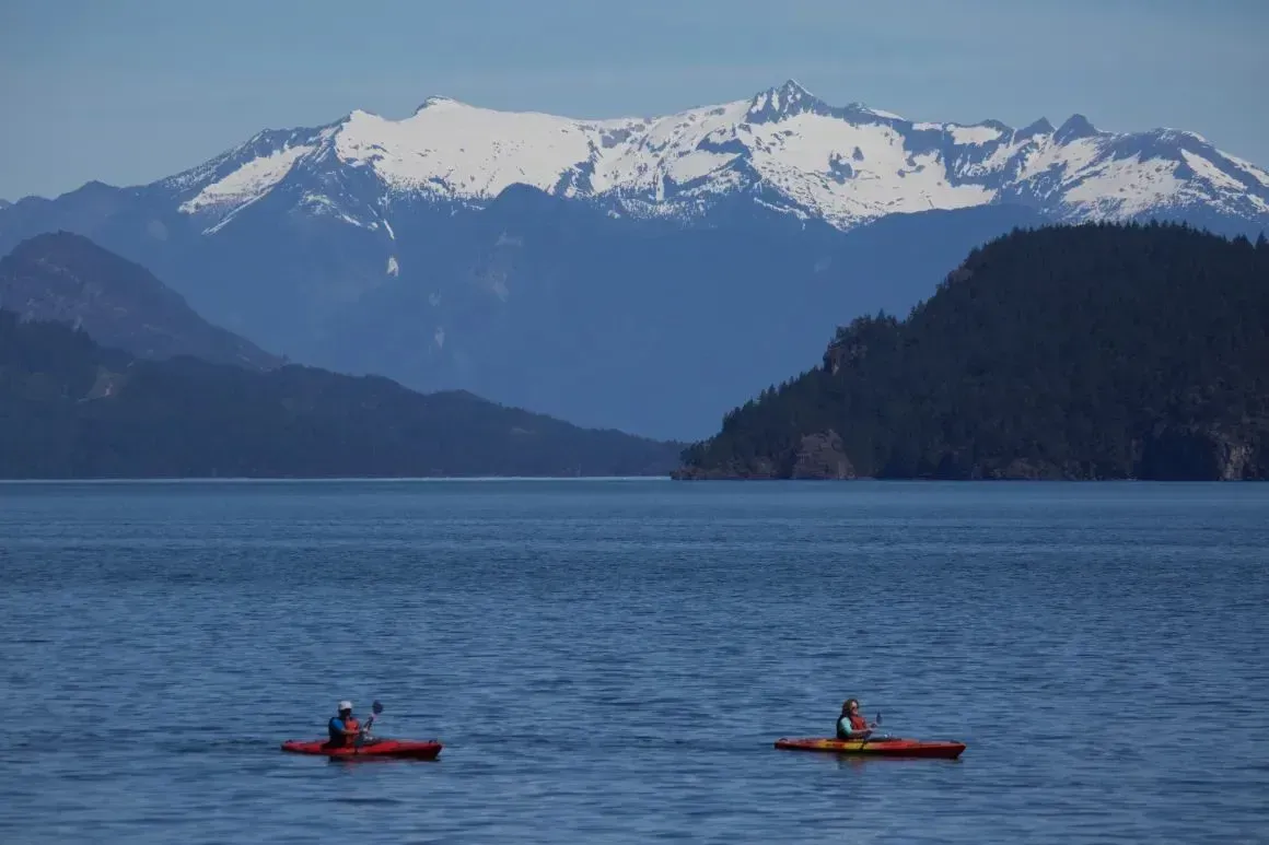 Two kayakers on calm blue water with forested hills and snowy mountains in the background