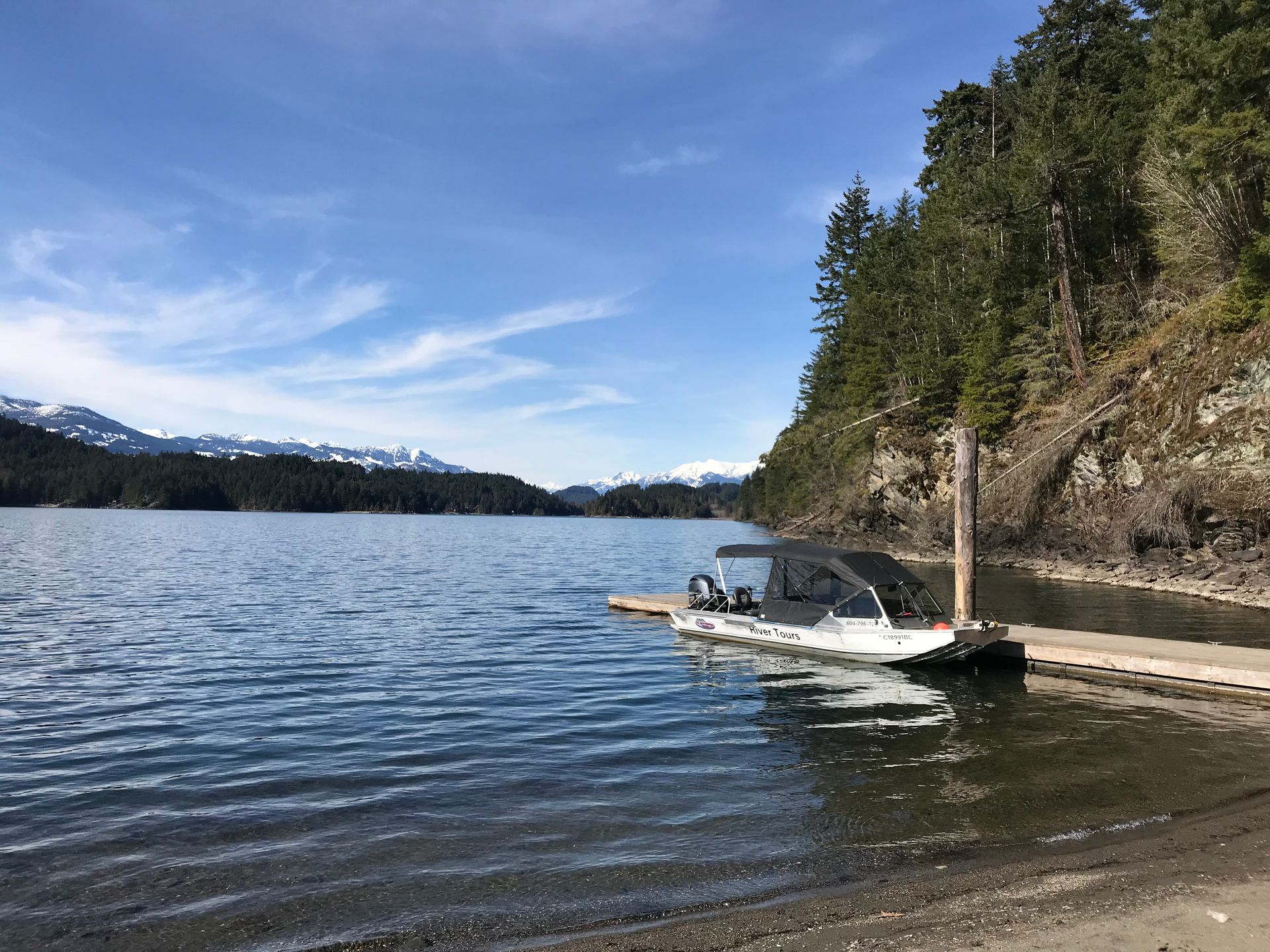 Small white motorboat at a lakeshore beside a rocky, forested cliff under a blue sky