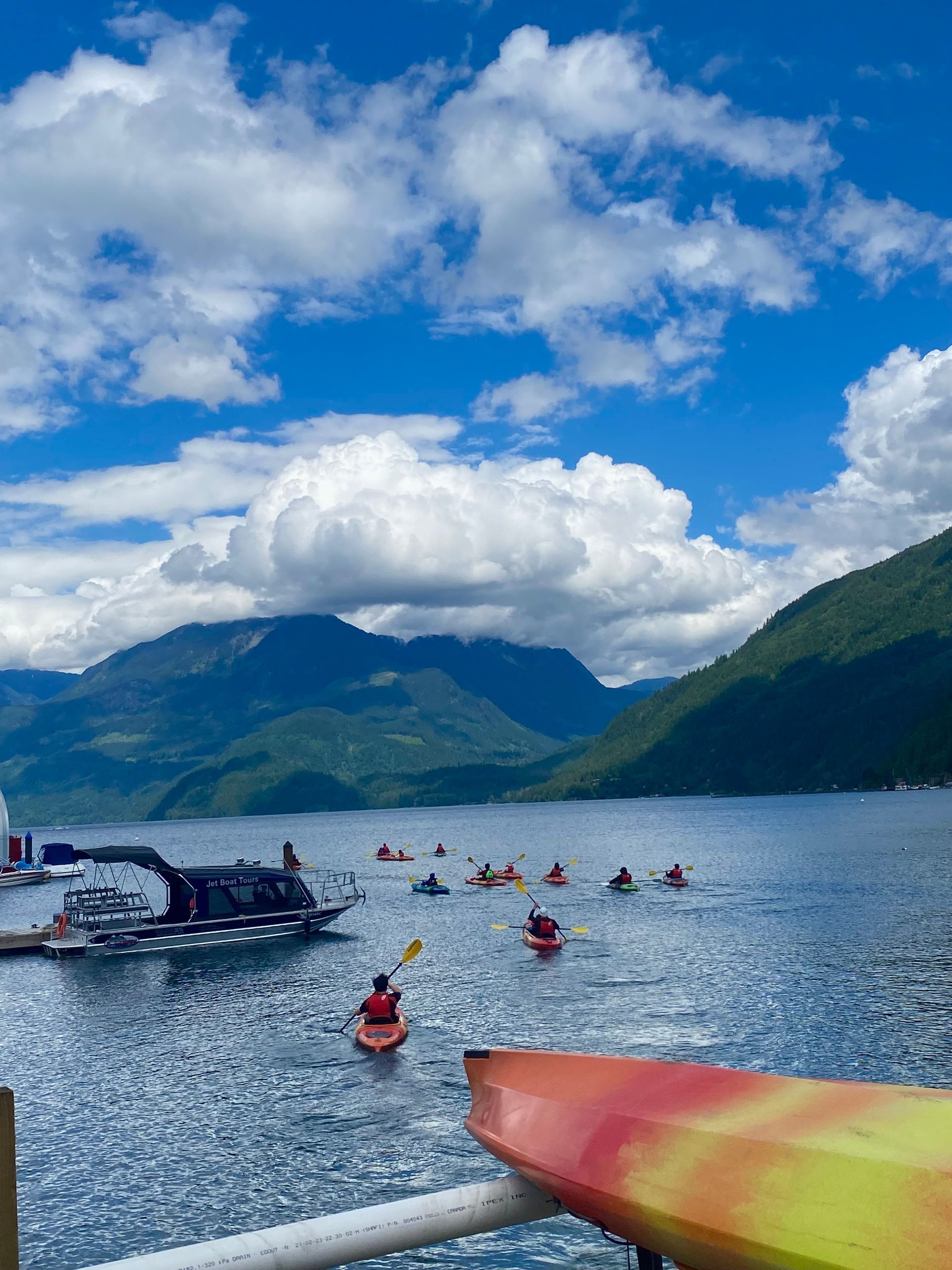 Kayakers on a blue lake with mountains and clouds; colorful kayak bow in the foreground
