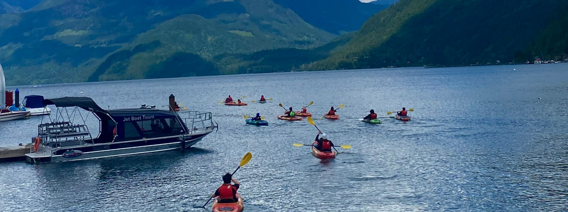 Kayakers in red vests paddle across a calm blue lake with a motorboat and green mountains in the background