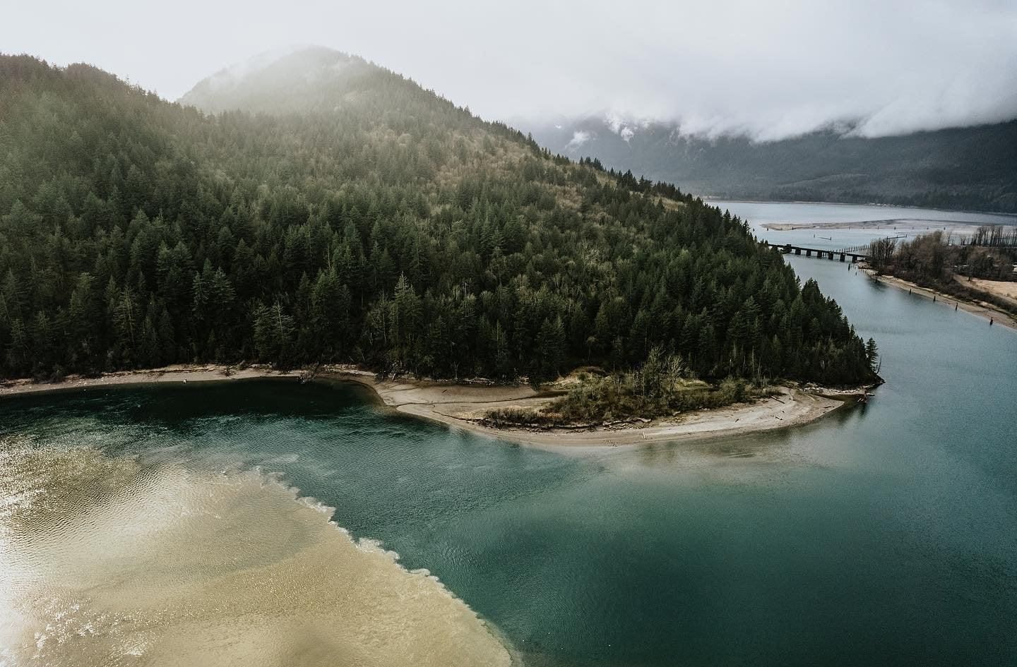 Foggy aerial view of a forested peninsula curving into calm blue water and sandy shoreline
