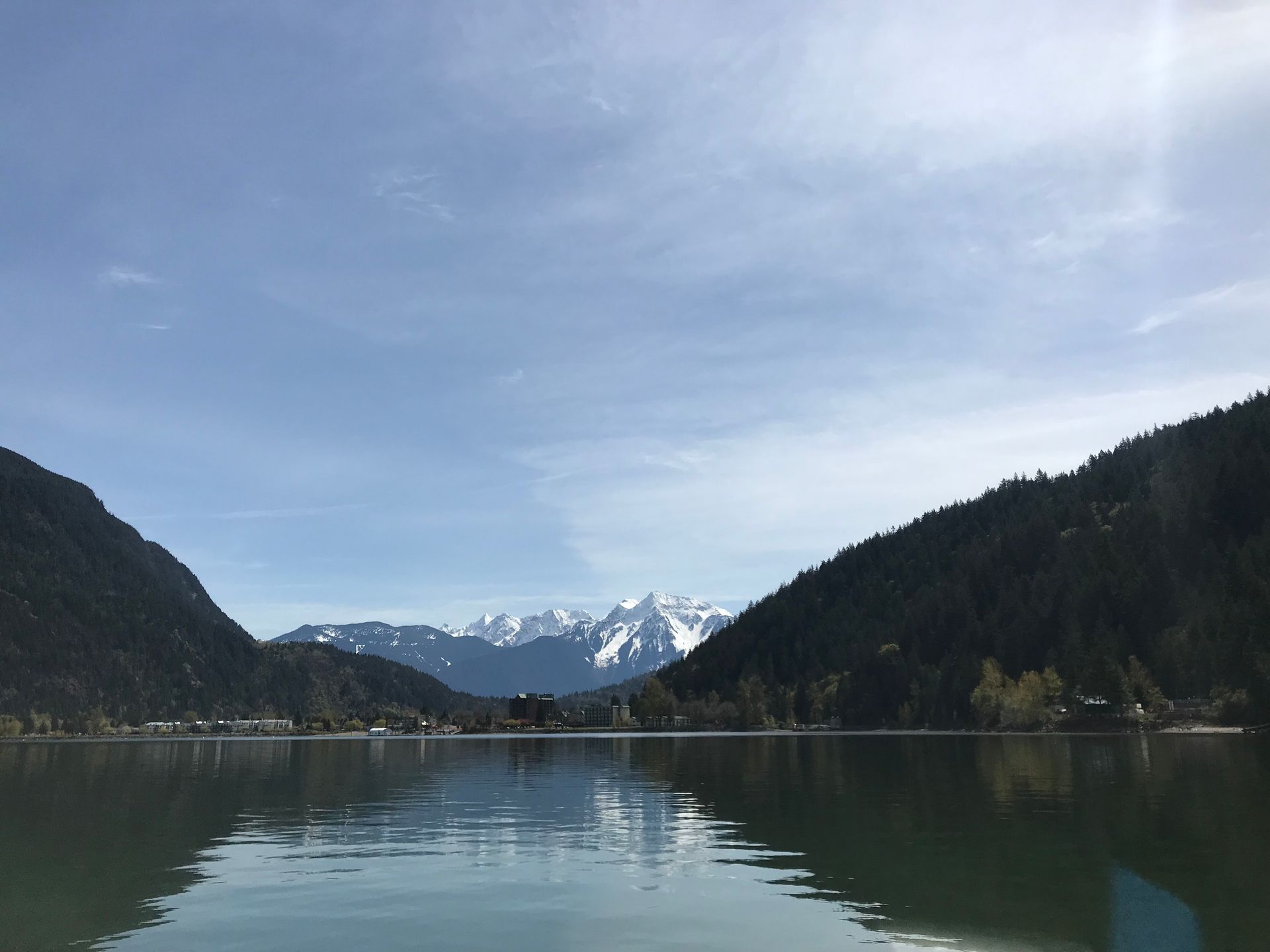 Snowy mountains reflected in a calm lake between forested hills under a cloudy sky