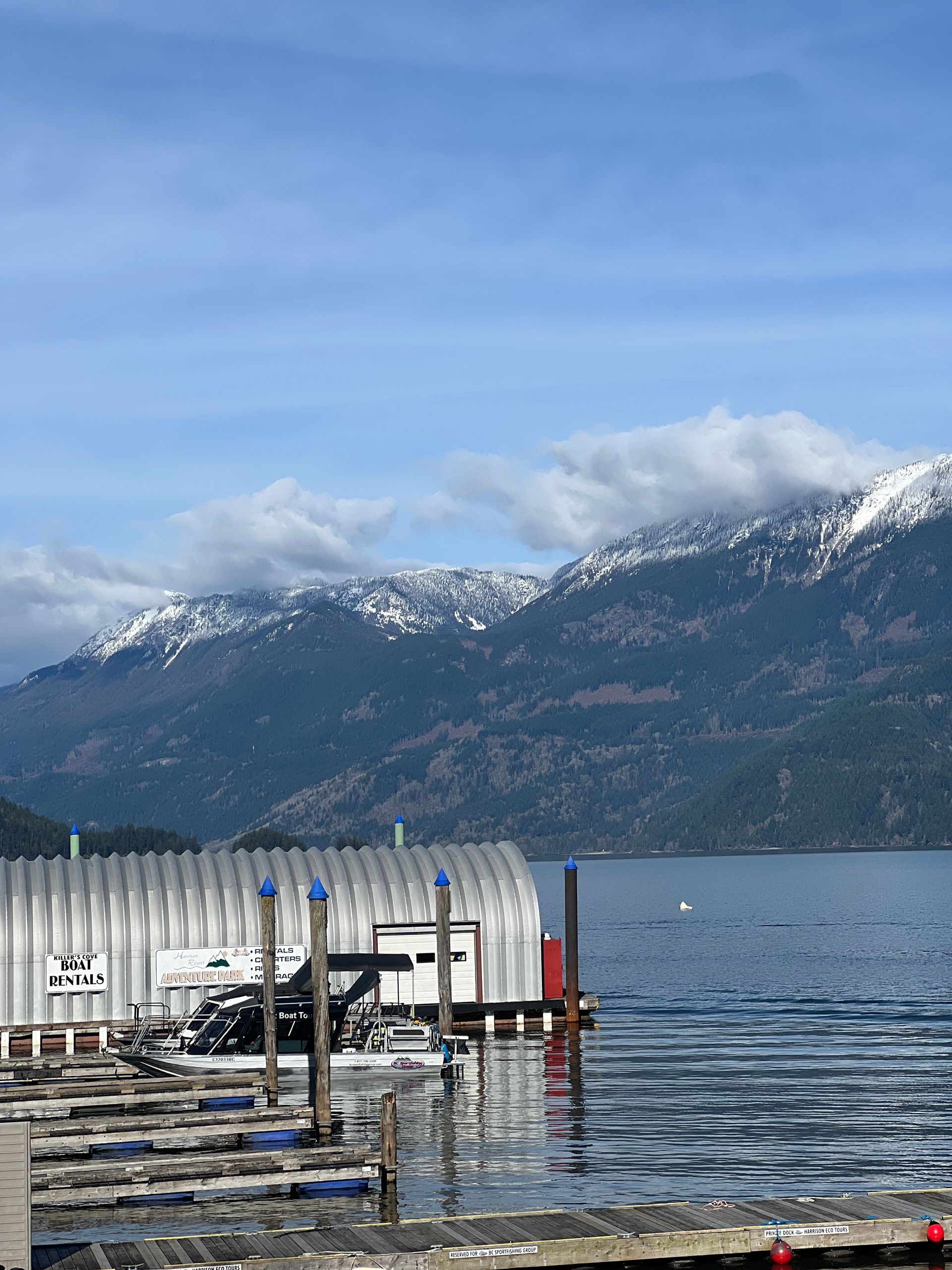 Snowy mountains beyond a calm lake, with a small dock in the foreground under a blue sky.