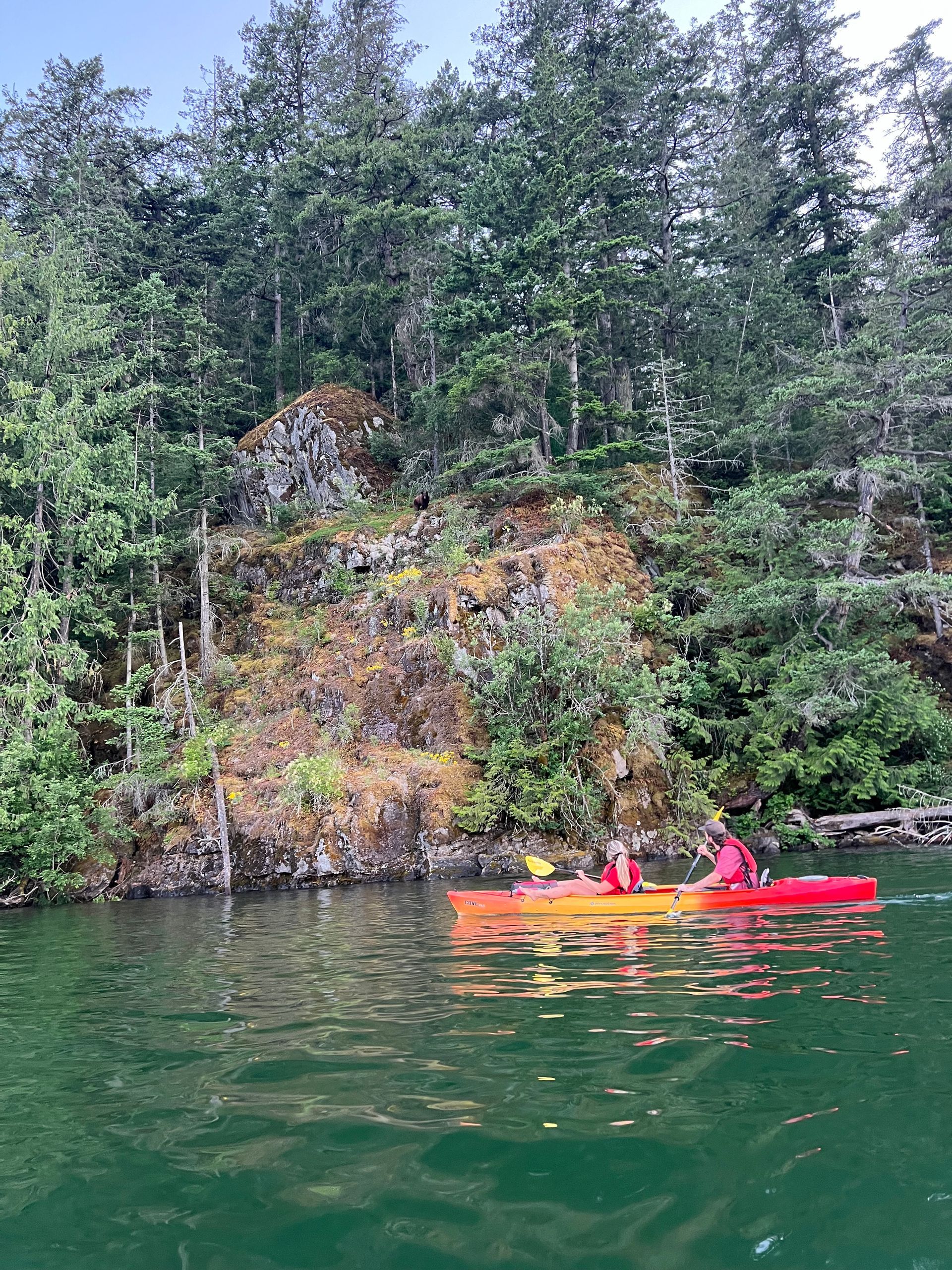 Two people in a red kayak on a green lake beside a rocky, forested cliff