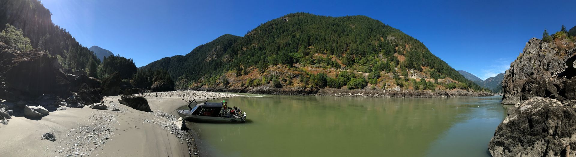 Rocky coastal cove with calm green water, sandy shore, and lush hills under a clear blue sky