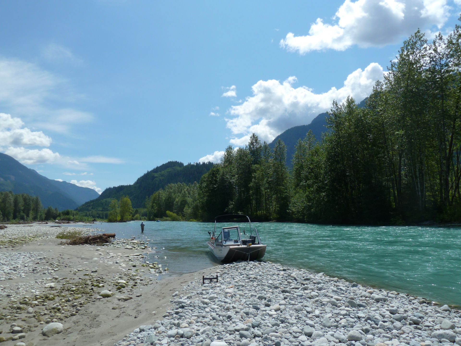 A small boat on a rocky riverbank beside turquoise water, with mountains and trees under a blue sky.
