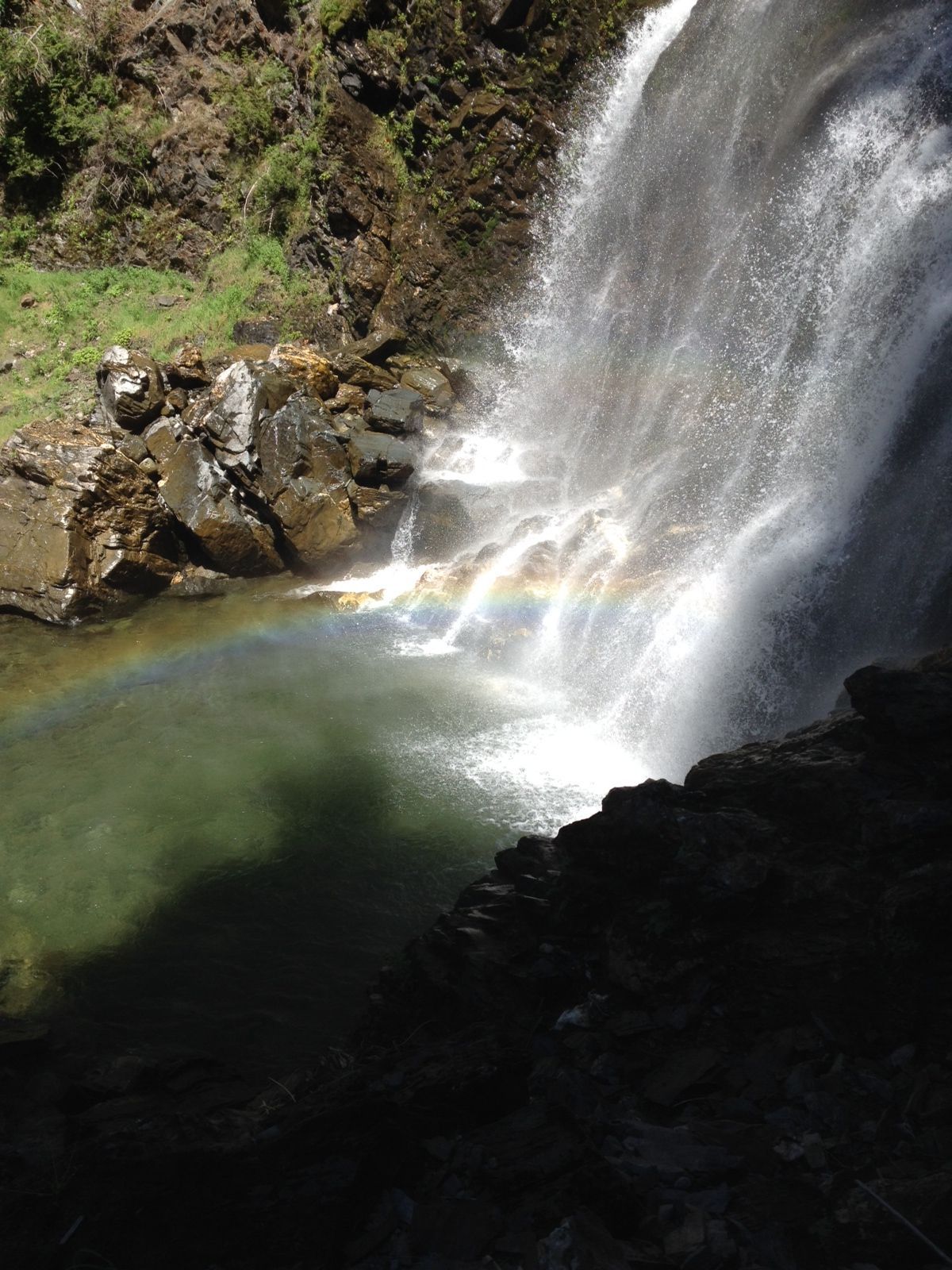 Waterfall cascading into a clear pool, with rainbow-like spray and rocky, shadowed surroundings.