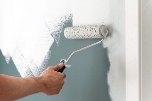 Person using a paint roller to apply white paint to a gray wall indoors.