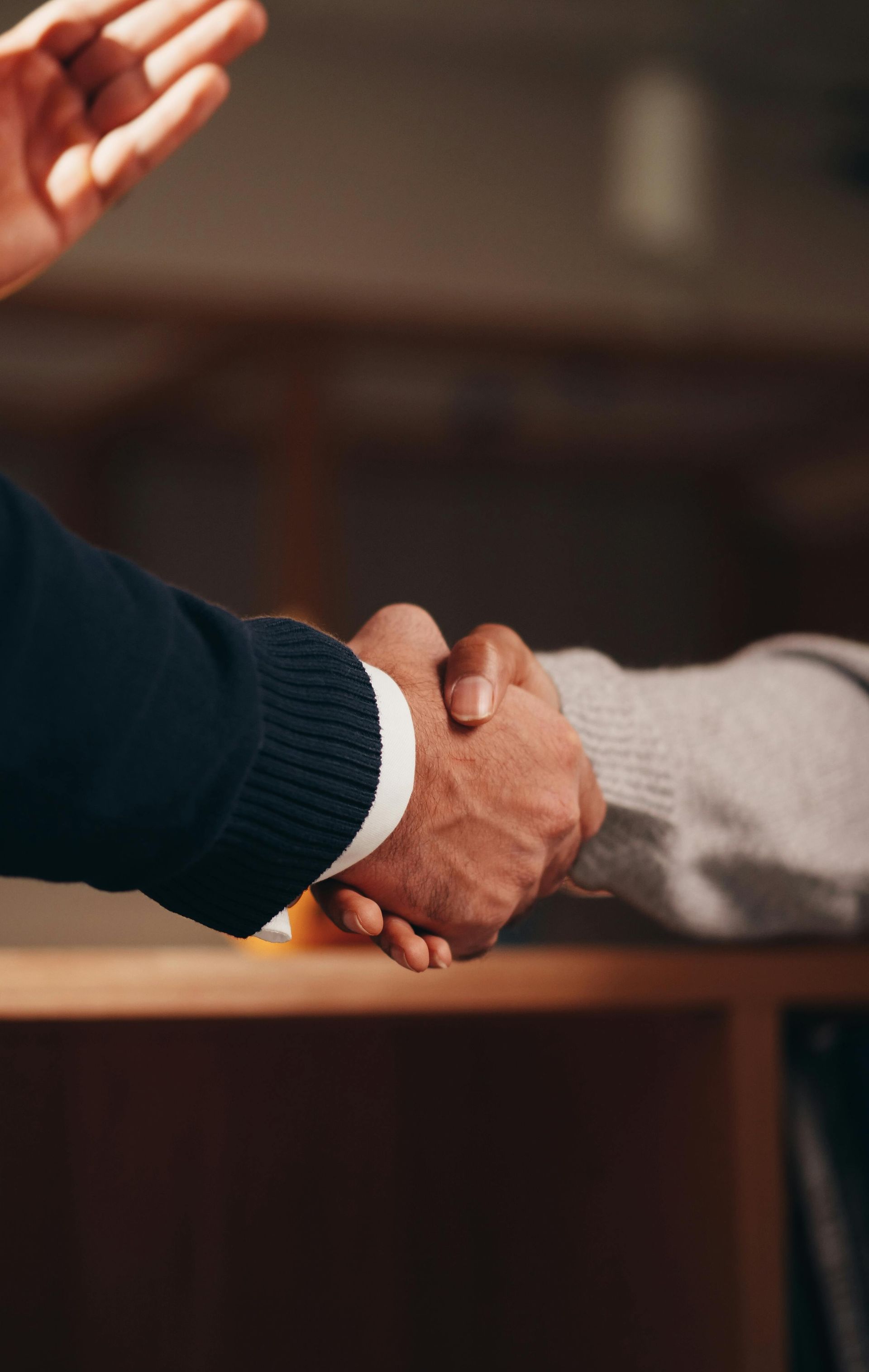 Two people shaking hands, one hand is reaching upward. Dark skin, and white and gray sleeves.