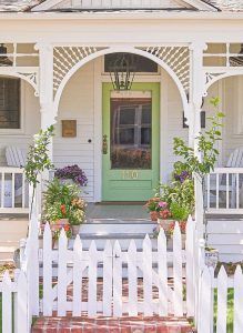 White cottage with green door, picket fence, and potted flowers on the porch.