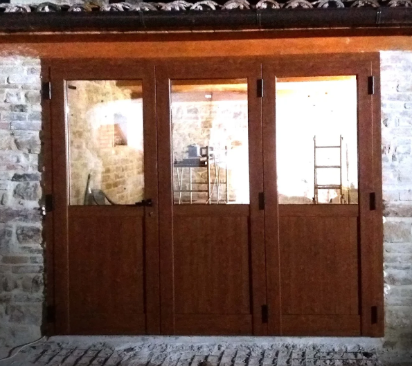 Doppie porte di legno con pannelli di vetro incastonate in un muro di pietra, che lasciano intravedere un interno illuminato con impalcature.