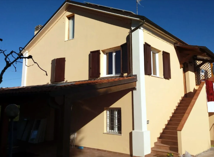 Casa beige a due piani con persiane marroni, portico coperto e scala esterna illuminata dal sole.
