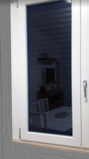Finestra con cornice bianca, tenda scura e un debole riflesso di una stanza interna.