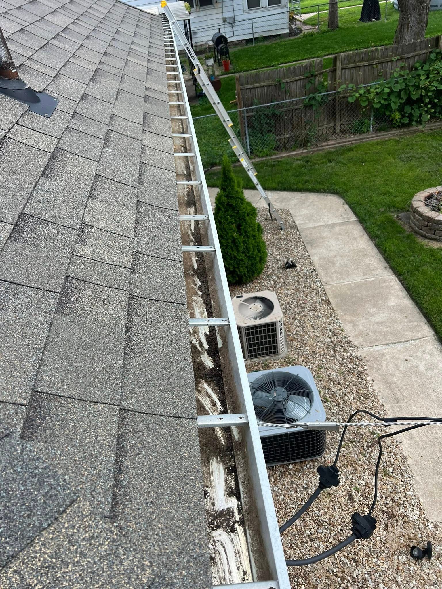 A view from a roof showing gutters equipped with metal mounting brackets, with a ladder leaning against the house below.