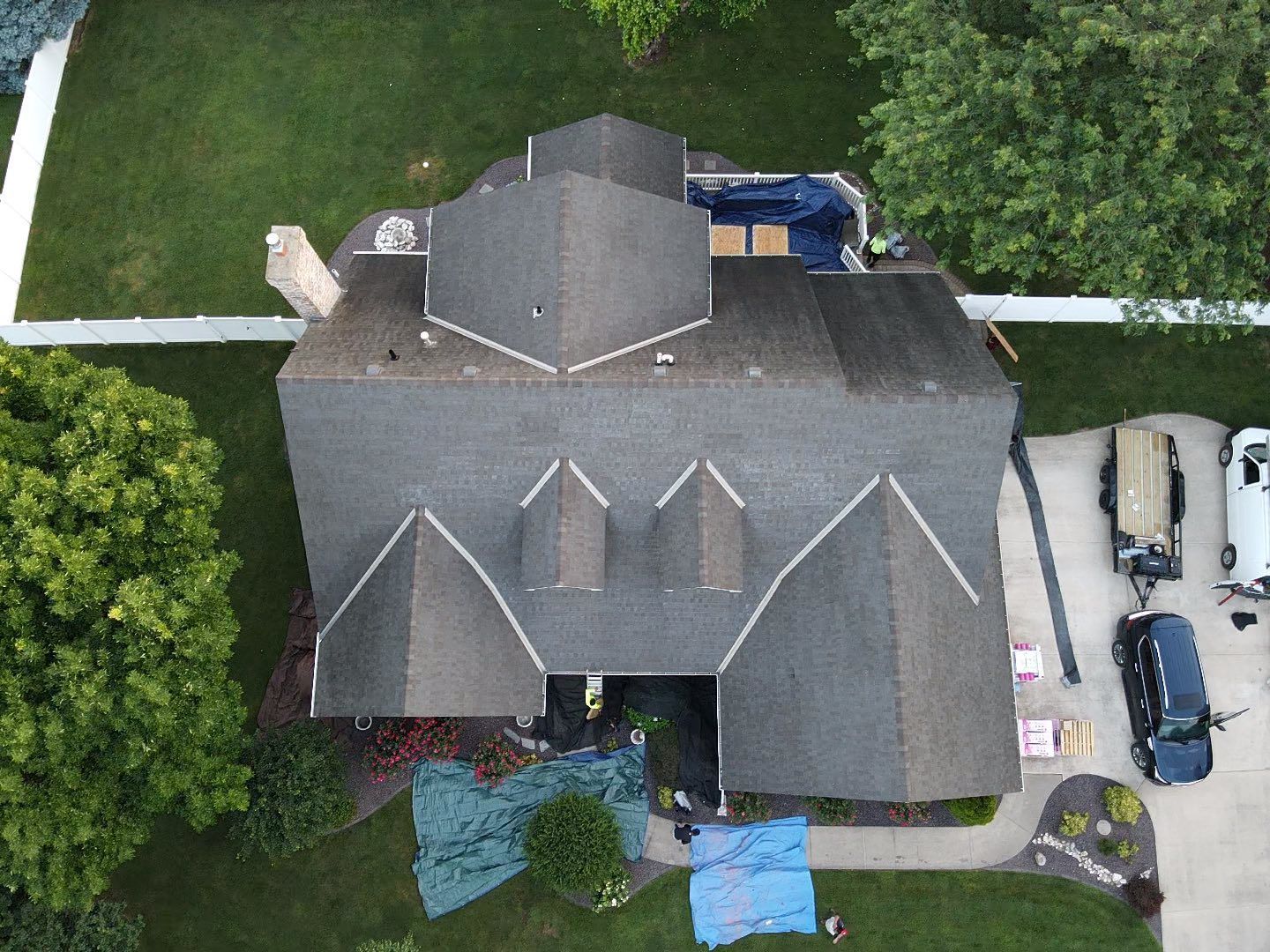 Aerial view of a gray-roofed house with surrounding yard, trees, and parked cars by a driveway.